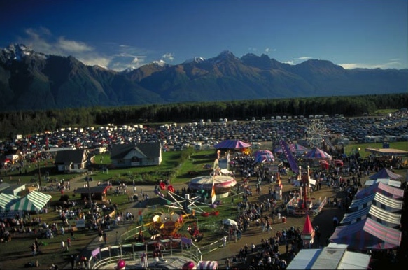 The Alaska state fair with rides and tents in the foreground and a full, expansive parking lot behind and jagged peaks behind Alaska State Fair