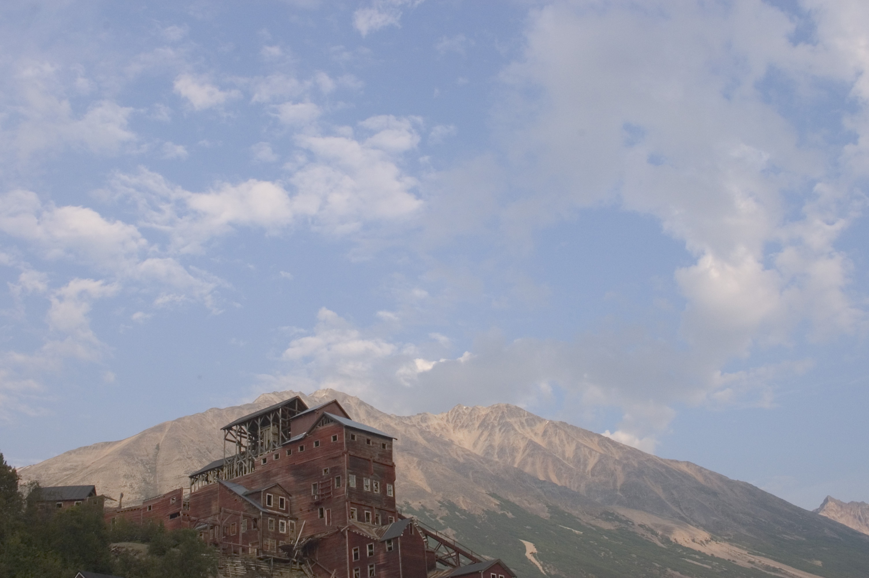 The ruins of the 1900-era Kennicott copper mine in Wrangell-St Elias National Park. Photo courtesy State of Alaska Tourism / Matt Hage. Kennicott copper mine with mountains behind on a sunny day with scattered clouds